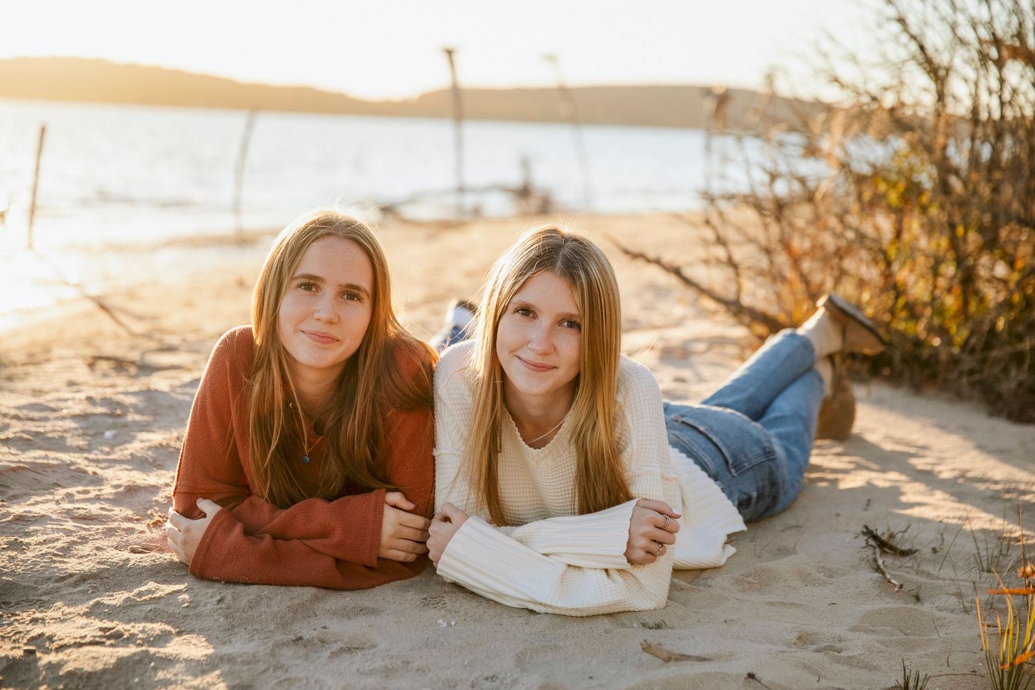 Two teenage girls lying on sandy beach at golden hour, wearing casual sweaters and jeans near a lake.