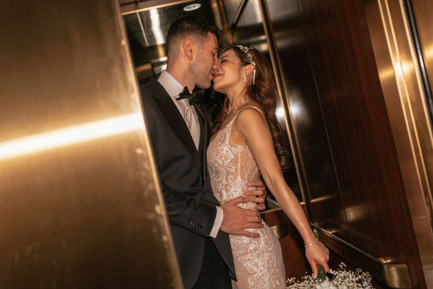 Bride and groom sharing a romantic moment in a golden elevator, dressed in wedding attire.