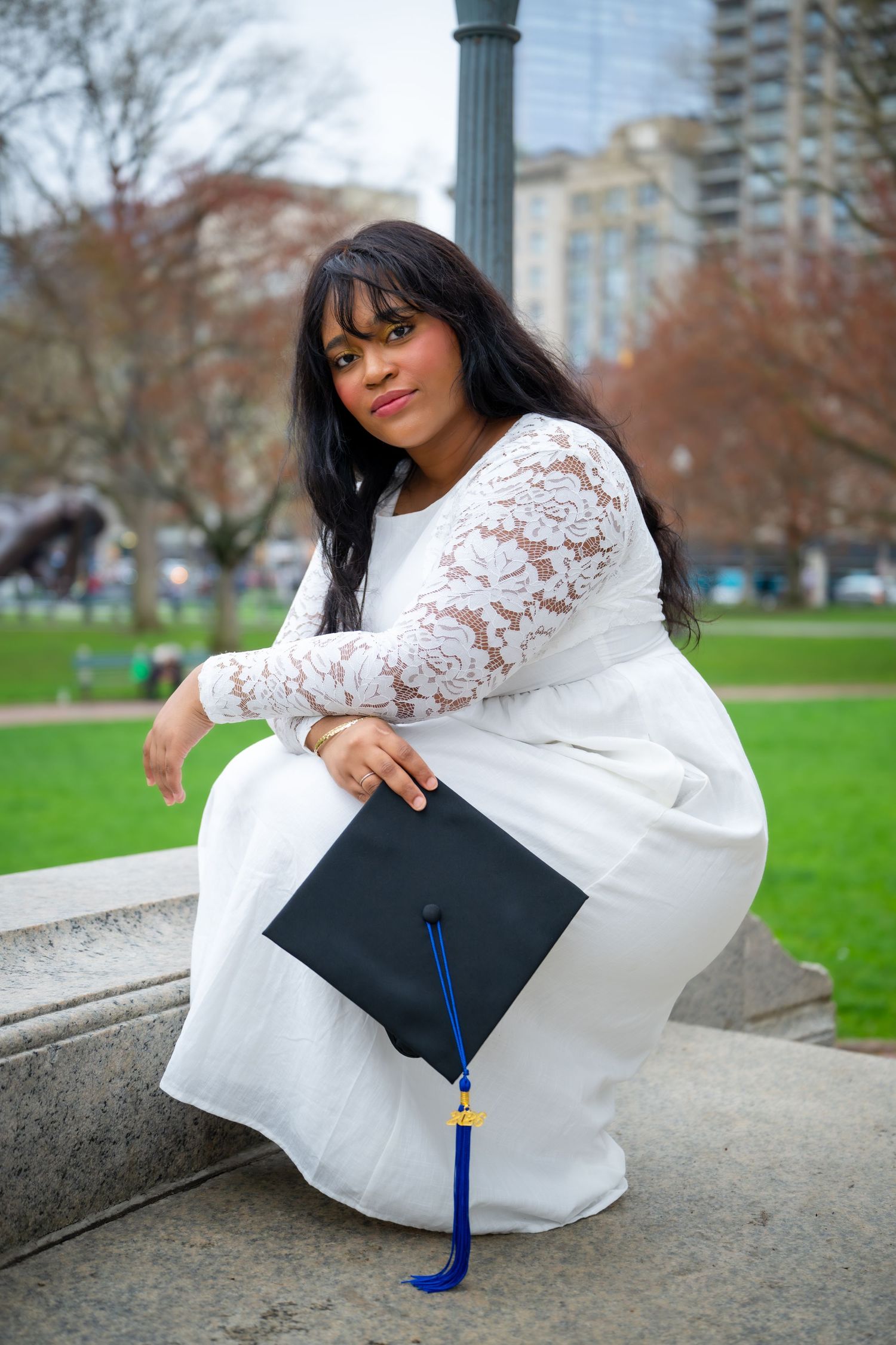 Young woman in white lace dress holding black graduation cap poses outdoors in urban park setting.