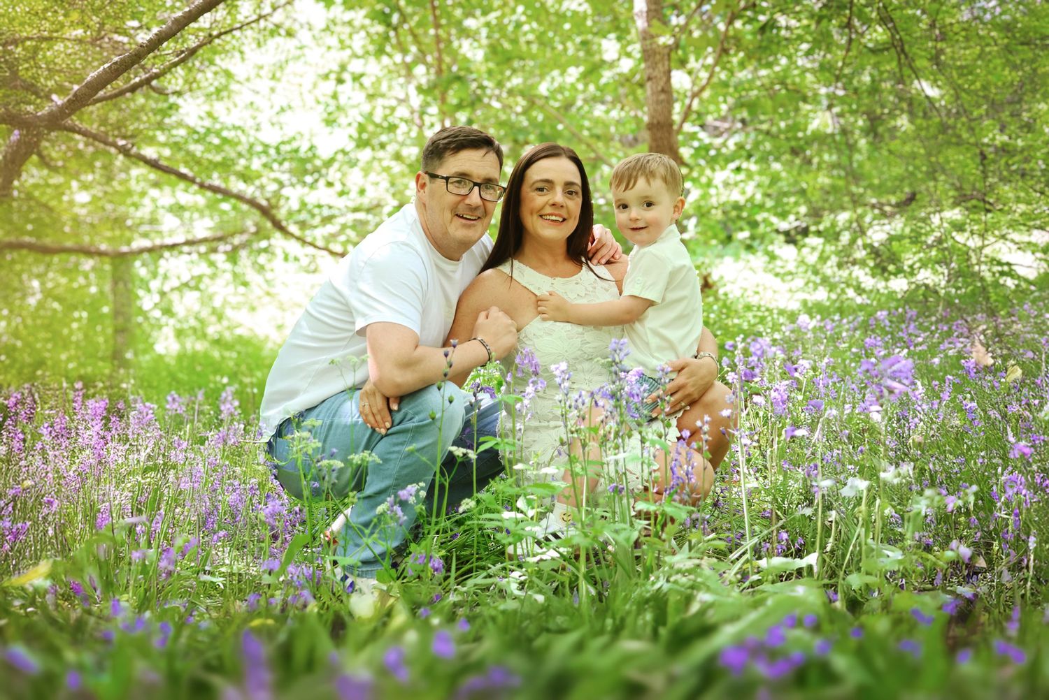 Family of three smiling among purple bluebells in a lush green woodland setting on a sunny day.