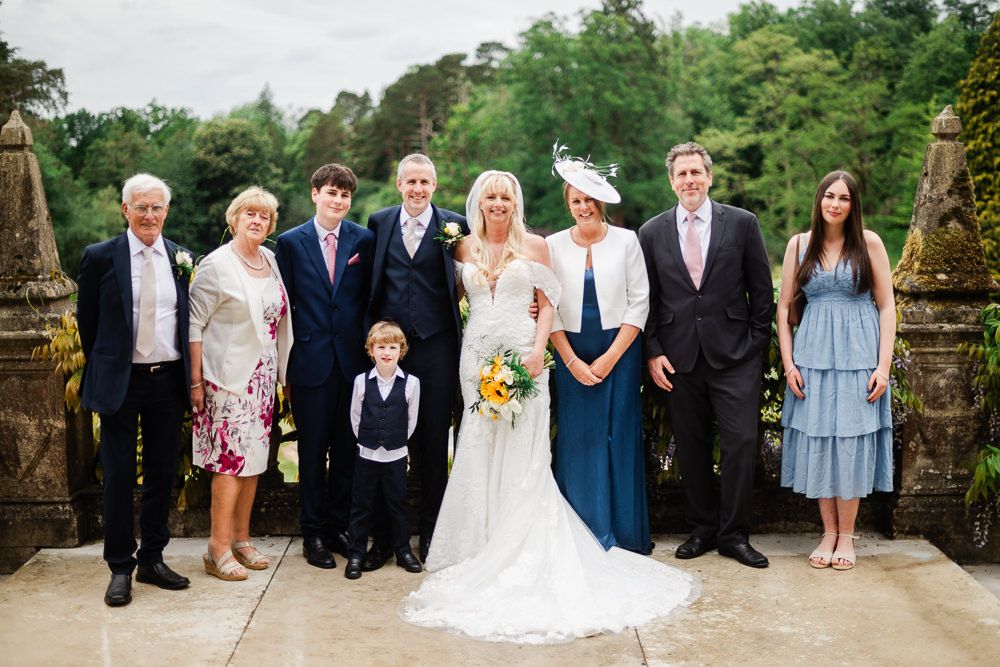 Wedding party posing outdoors at elegant venue, bride in white lace gown with bouquet surrounded by family in formal attire.