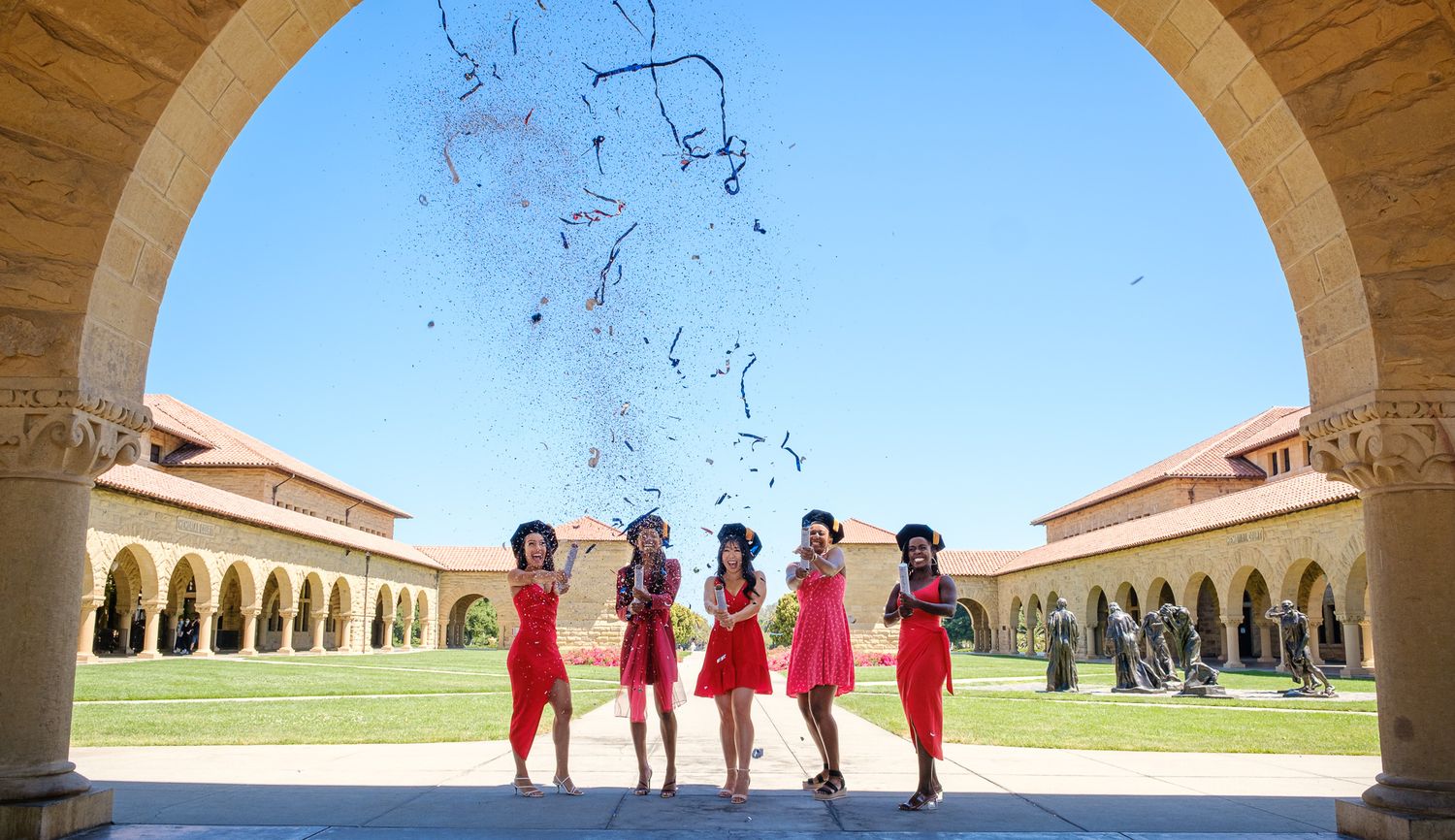 Five women in red dresses celebrate under an arch, tossing confetti in the air at Stanford University.