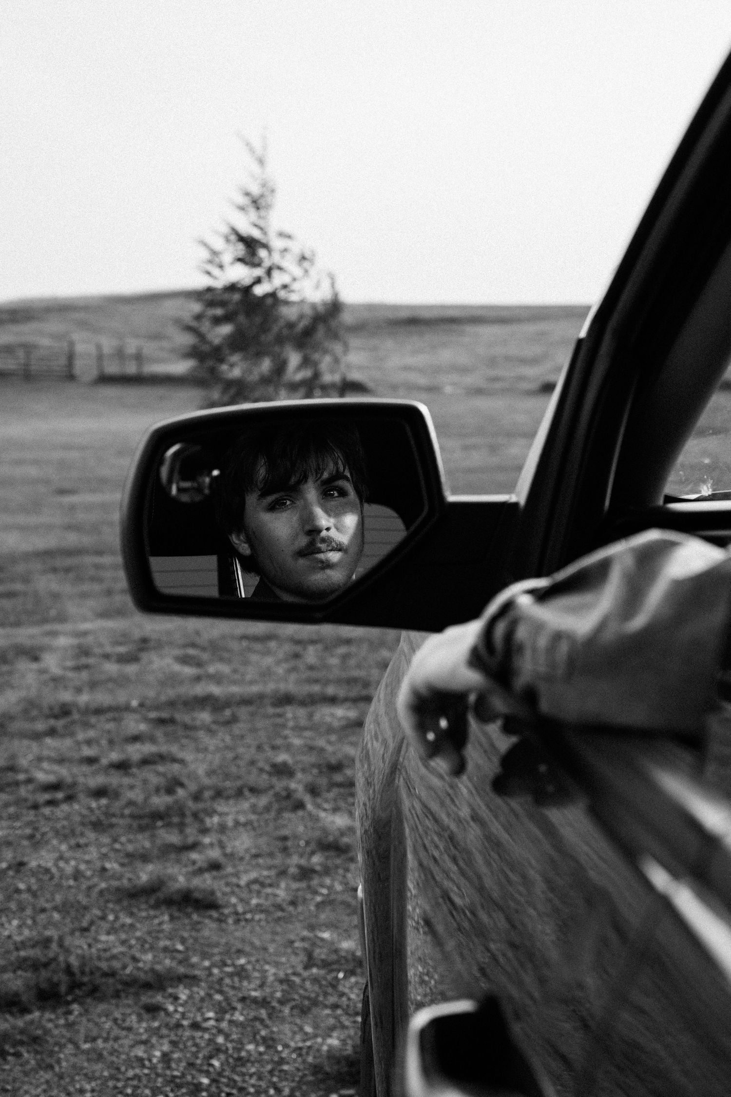 Black and white portrait of young woman reflected in car side mirror in rural countryside setting.
