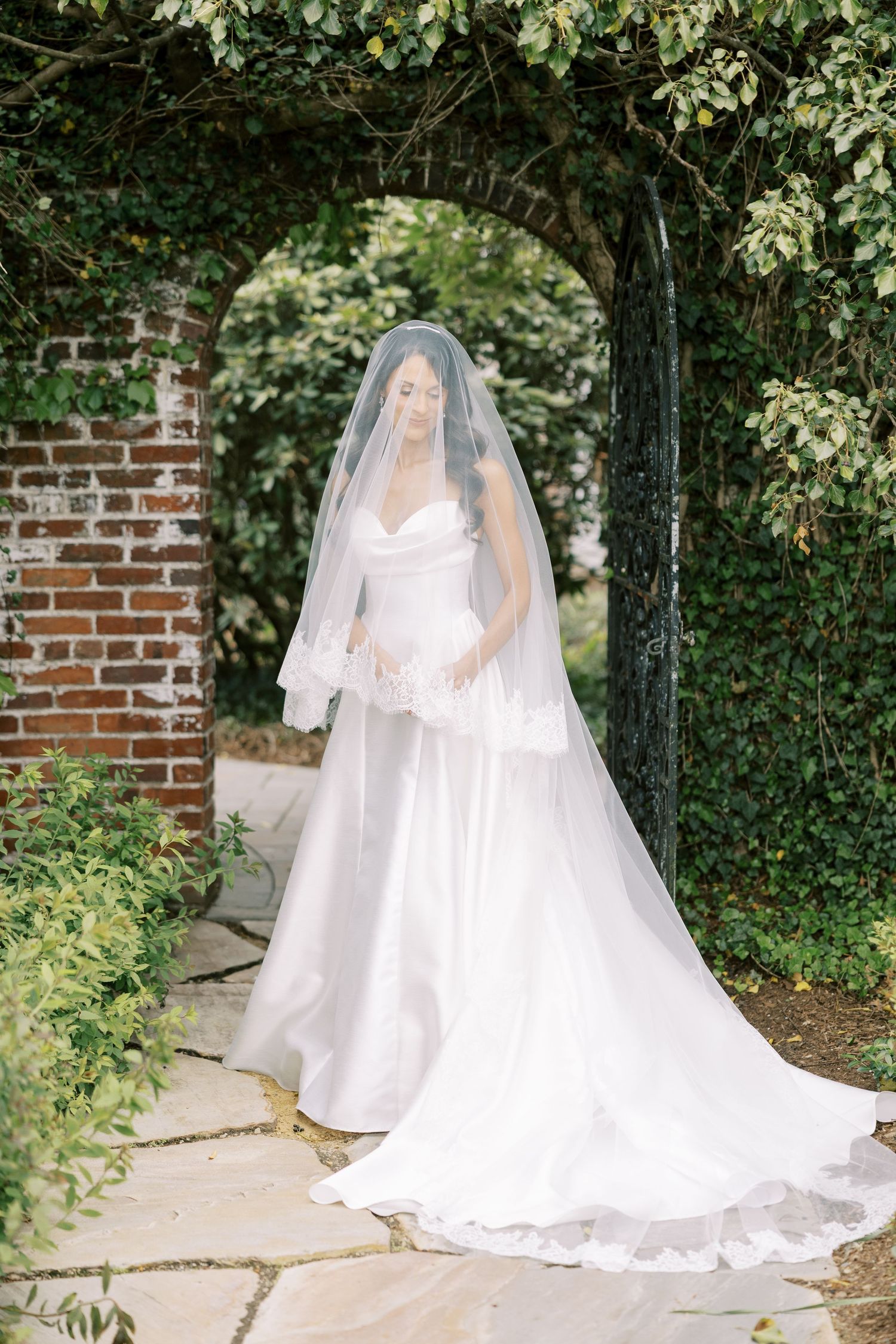 Bride in a white gown and lace veil stands under a brick archway surrounded by lush greenery on a stone path.