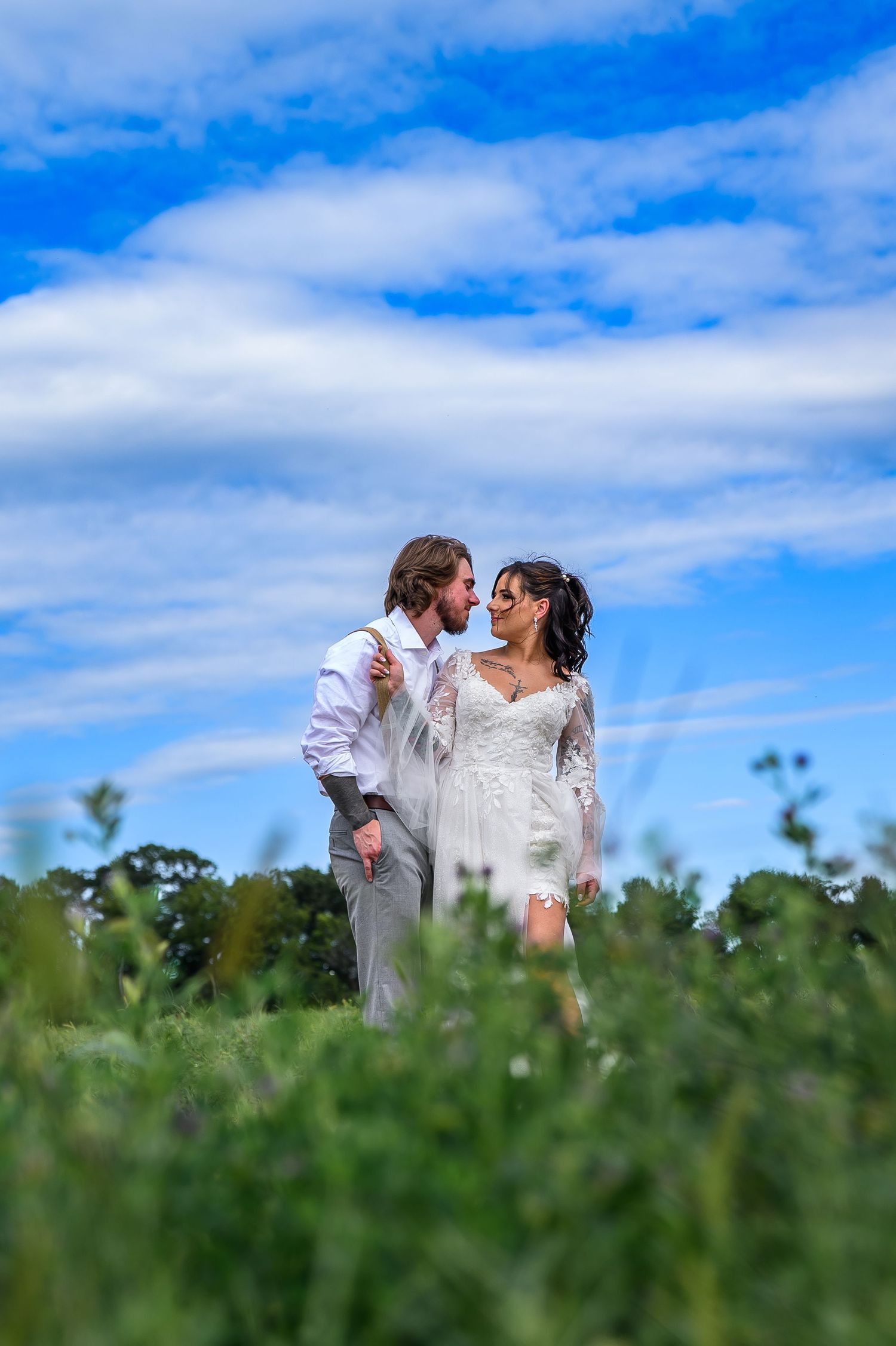 Bride and groom share a romantic moment in a lush green field under a bright blue sky on their wedding day.