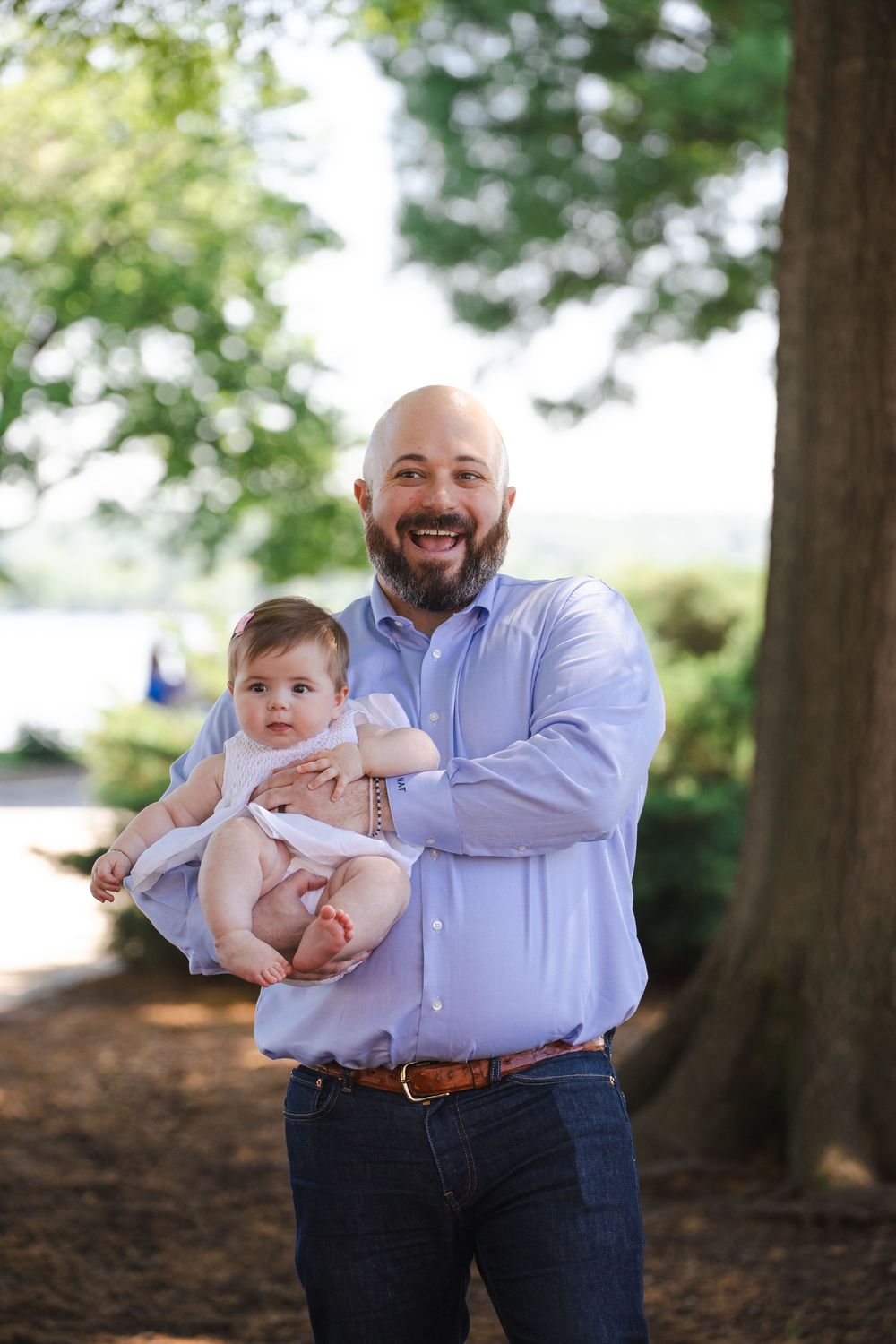 Smiling bald man with beard holding a baby outdoors among green trees, wearing a light blue shirt and jeans.
