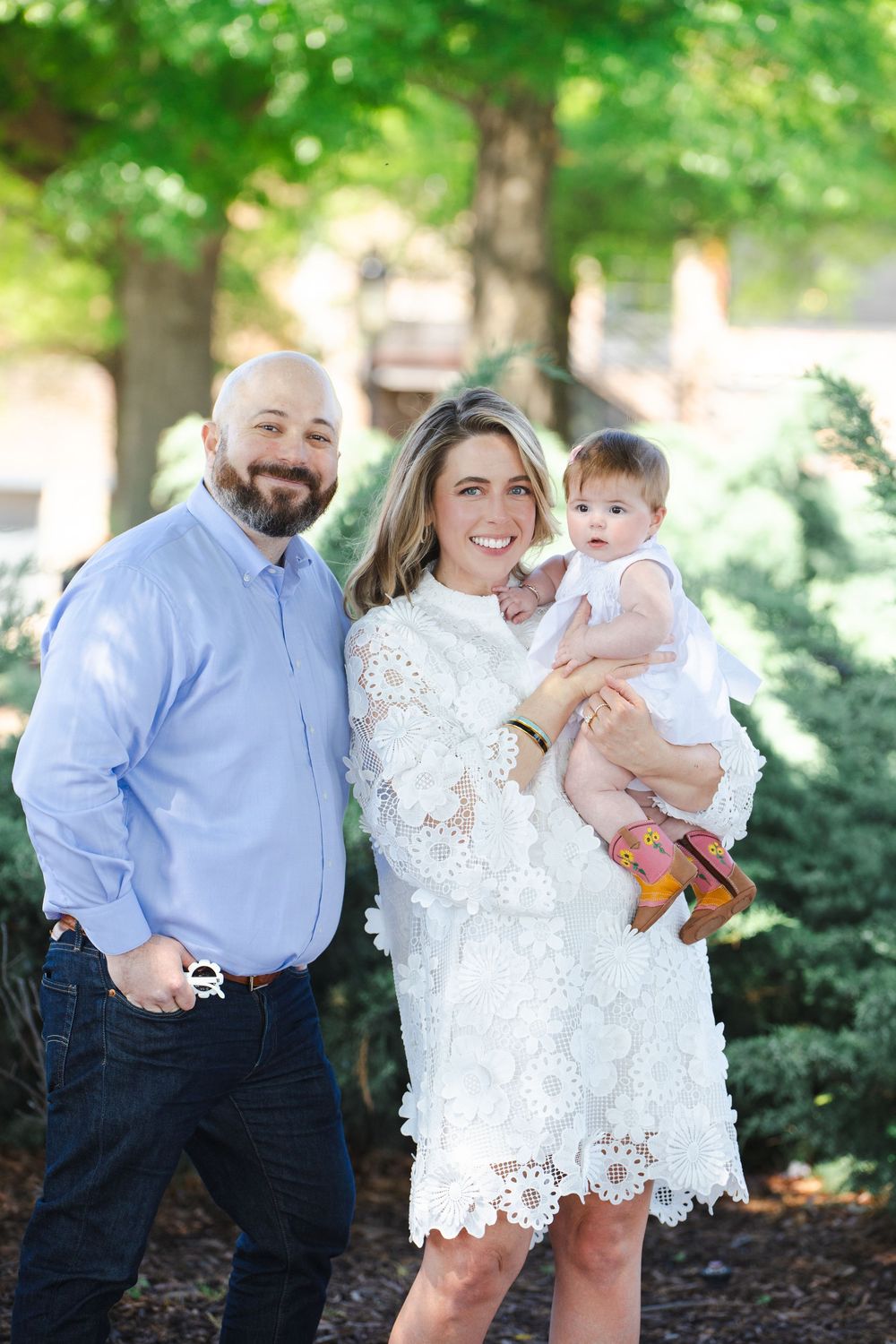 A smiling couple poses outdoors with their baby girl, the woman wearing a white lace dress while holding the child.
