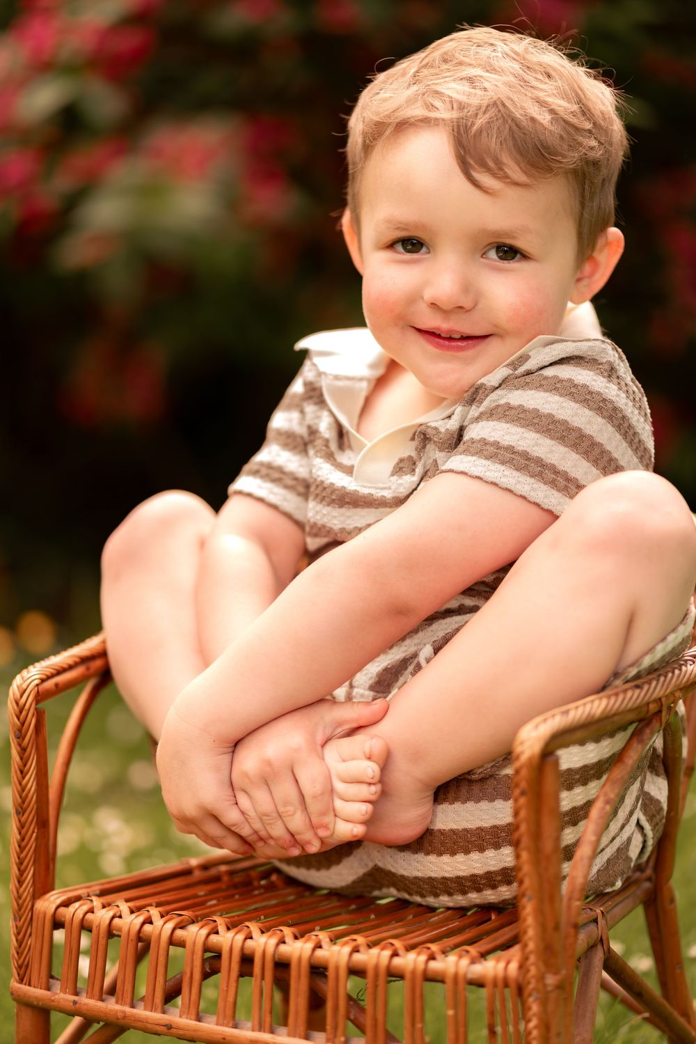 Young smiling boy in striped romper sitting cross-legged on wicker chair outdoors with pink flowers in background.