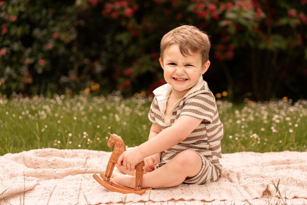 Smiling toddler boy in striped romper sitting on blanket outdoors holding wooden rocking horse toy in garden.