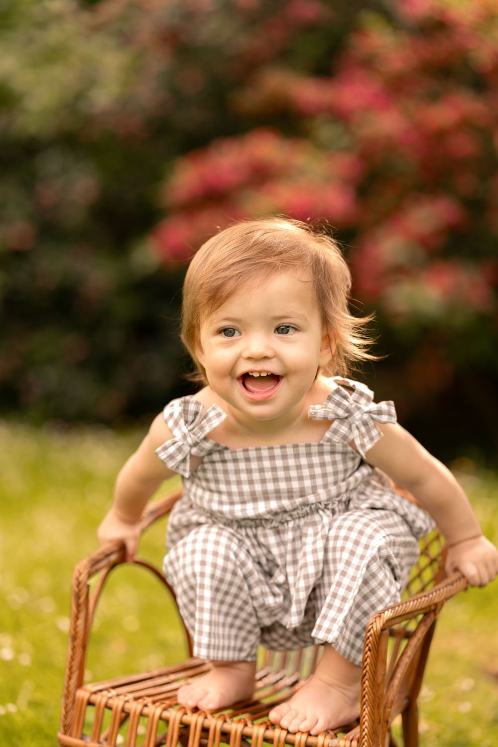 Happy baby girl in gingham romper sitting in wicker chair outdoors, smiling in a lush green garden.