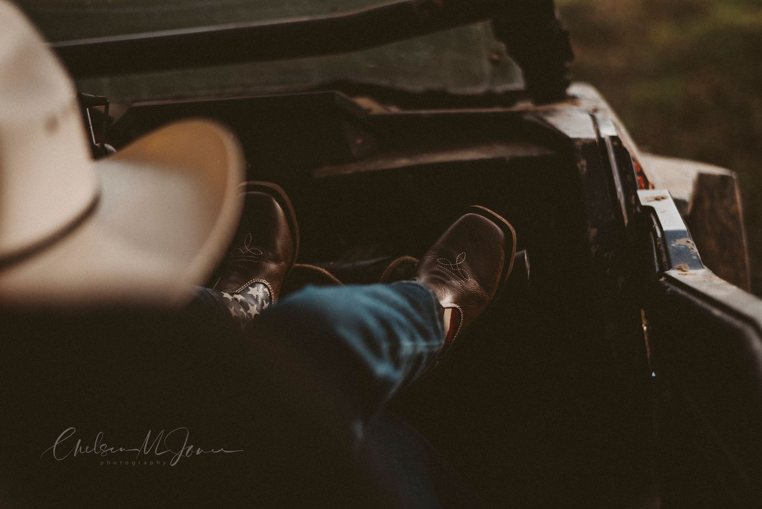 Person relaxing in a vintage car with brown leather shoes, jeans, and a cowboy hat in a moody outdoor setting.
