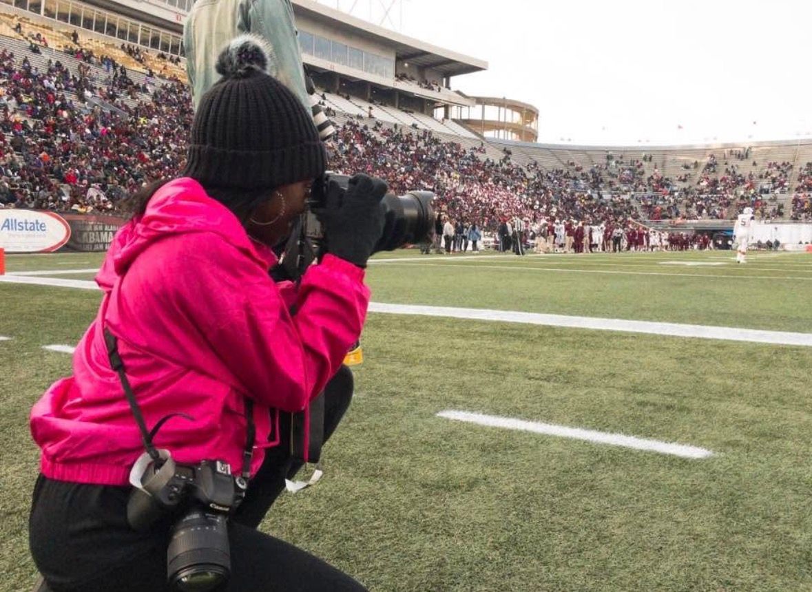 Photographer in pink jacket crouching on football field sideline capturing game action in packed stadium.