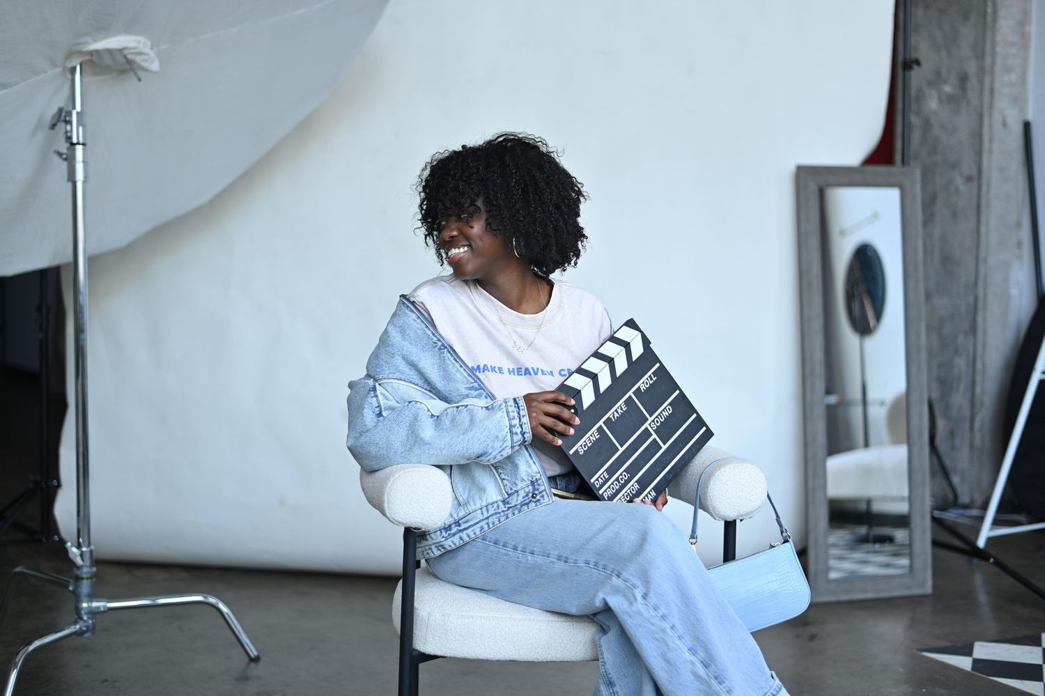 Young Black woman smiling in a photography studio, holding a film clapperboard while seated in a white chair.