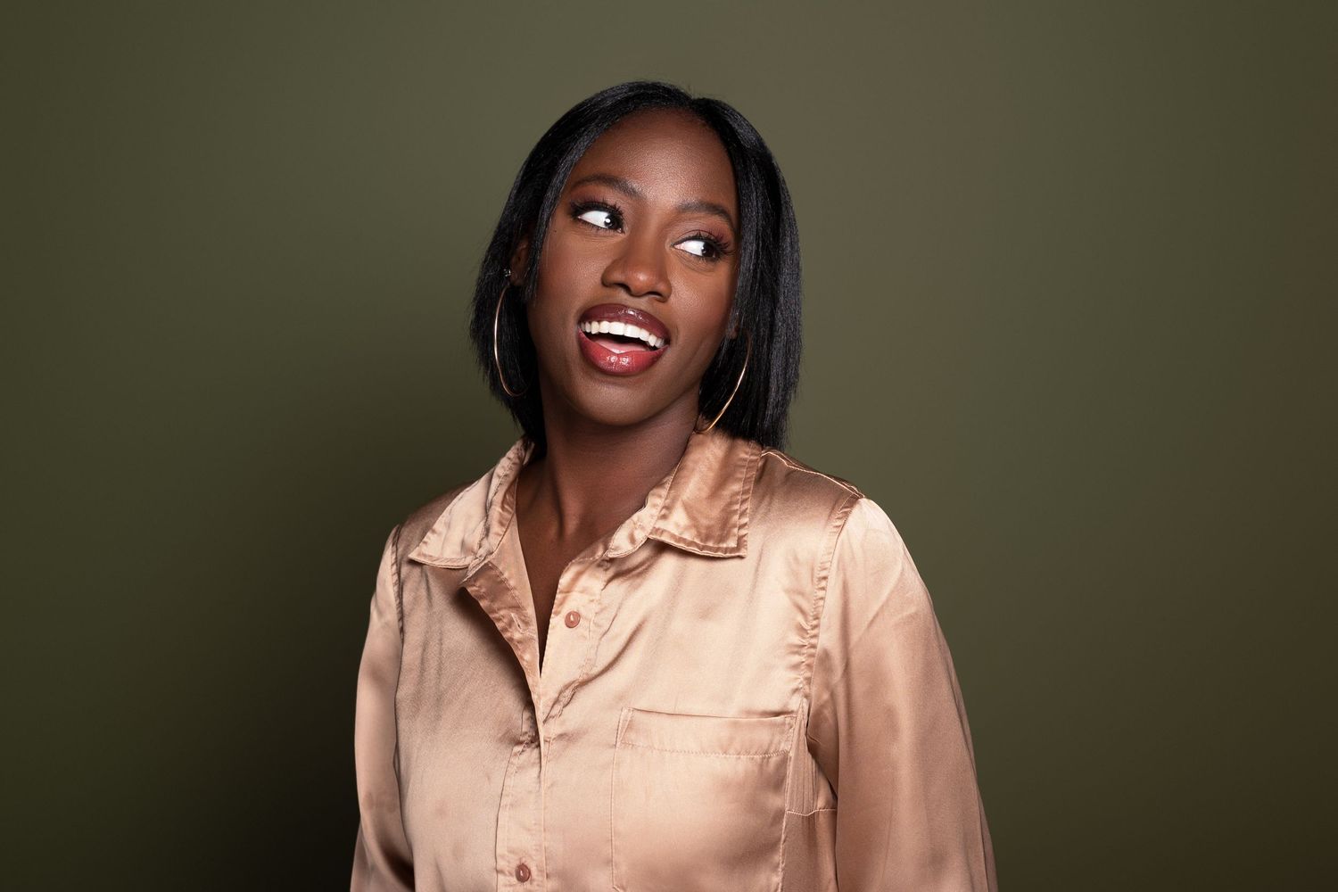 Young Black woman in a gold satin shirt smiling against an olive green background, wearing hoop earrings.