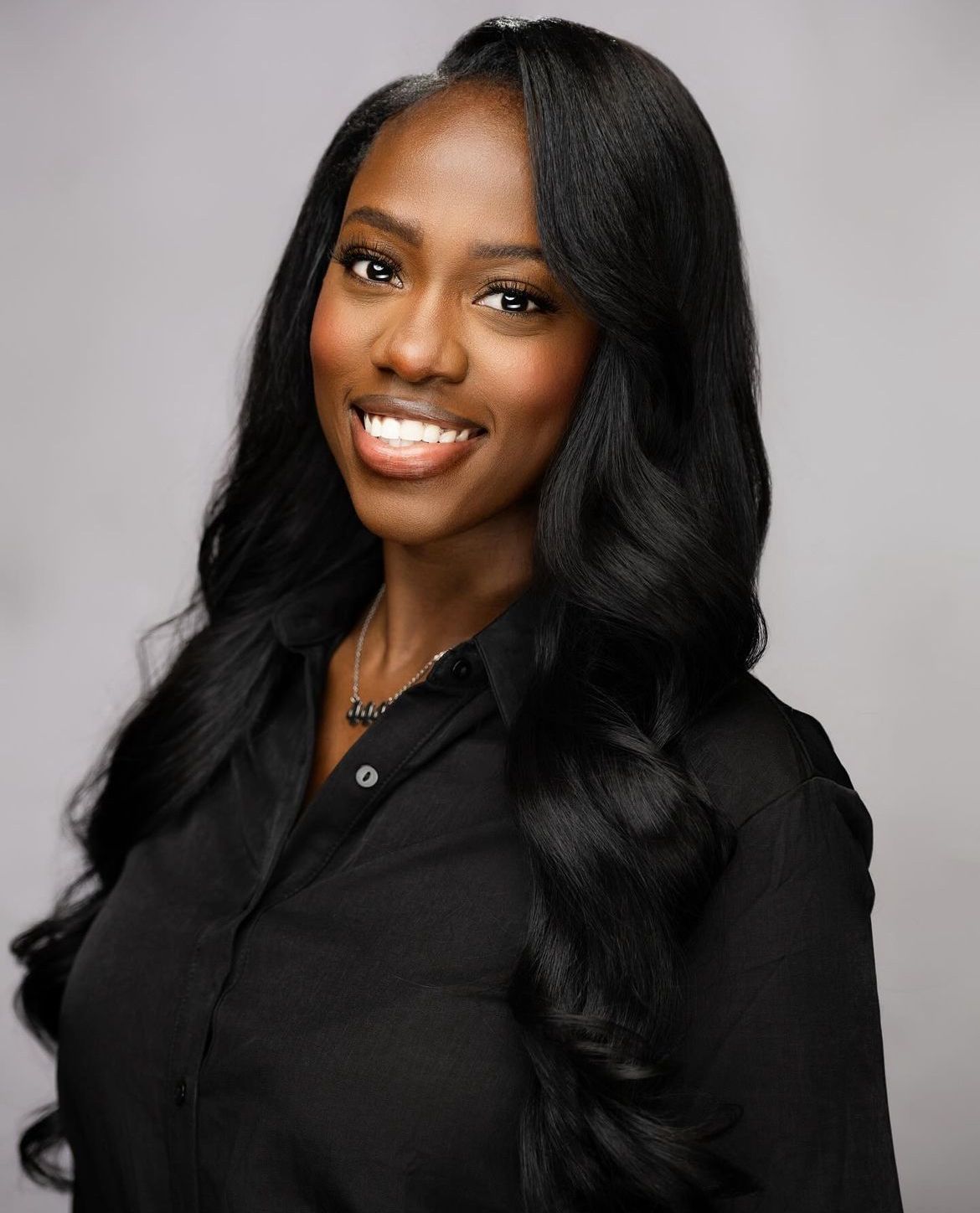 Professional headshot of a smiling woman with long black hair wearing a black blouse against a gray background.