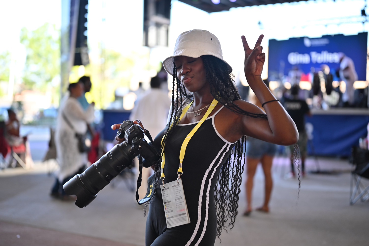 Woman photographer with braids, white hat, yellow lanyard, holding large camera lens, posing with peace sign outdoors.