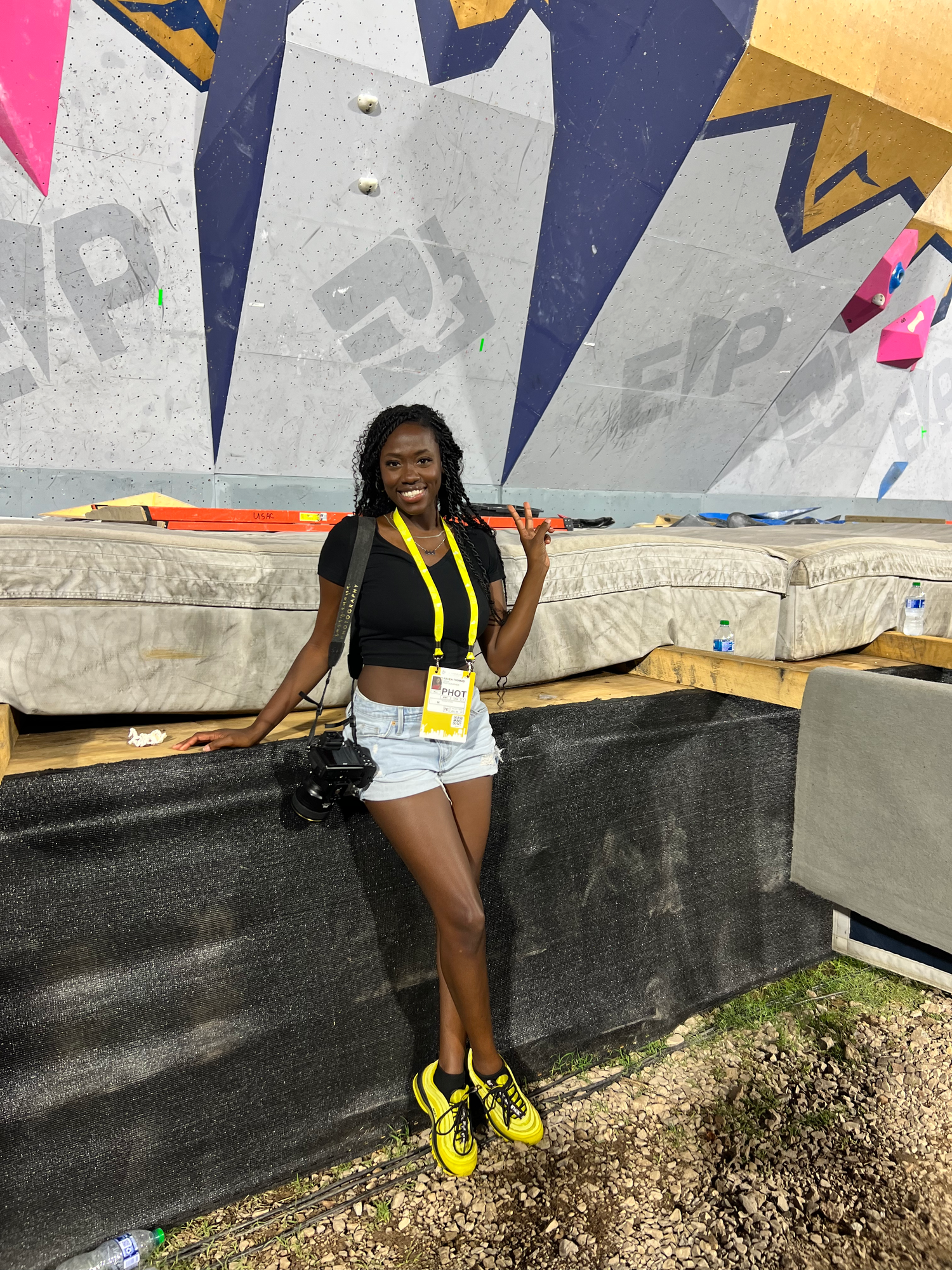 Young woman smiling in black crop top, denim shorts, yellow sneakers, lanyard, posing at indoor climbing wall facility.
