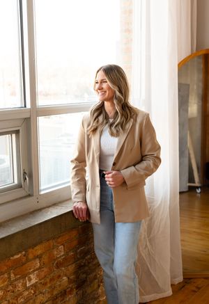 Woman in casual business attire smiles by bright window in modern apartment with exposed brick.