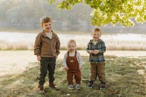 Children in matching brown overalls play together in a sunlit autumn meadow near a lake.
