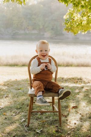 A smiling baby in brown overalls sits in a wooden chair outdoors during a sunny autumn day.