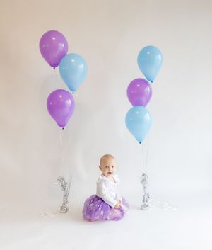 A series of blue and purple balloons floating against a white background with a baby in a tutu dress.