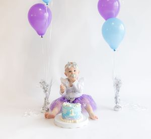 A baby in a blue and purple tutu dress sits on the floor with purple and blue balloons for a cake smash photo session.