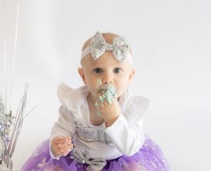 Baby in white and purple tutu dress with silver bow hair accessory eating cake during first birthday celebration.