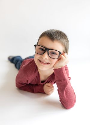 Child wearing glasses lies on white floor with head propped up on hand, showing a playful expression.