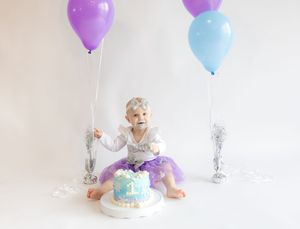 Purple and blue balloons float above a white cake in a minimalist studio setting.