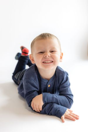A young child wearing a navy blue sweater lies on their stomach on a white background, smiling brightly at the camera.