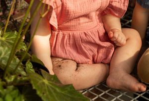 Small child in a pink romper sits outdoors near green foliage with bare feet and legs visible.