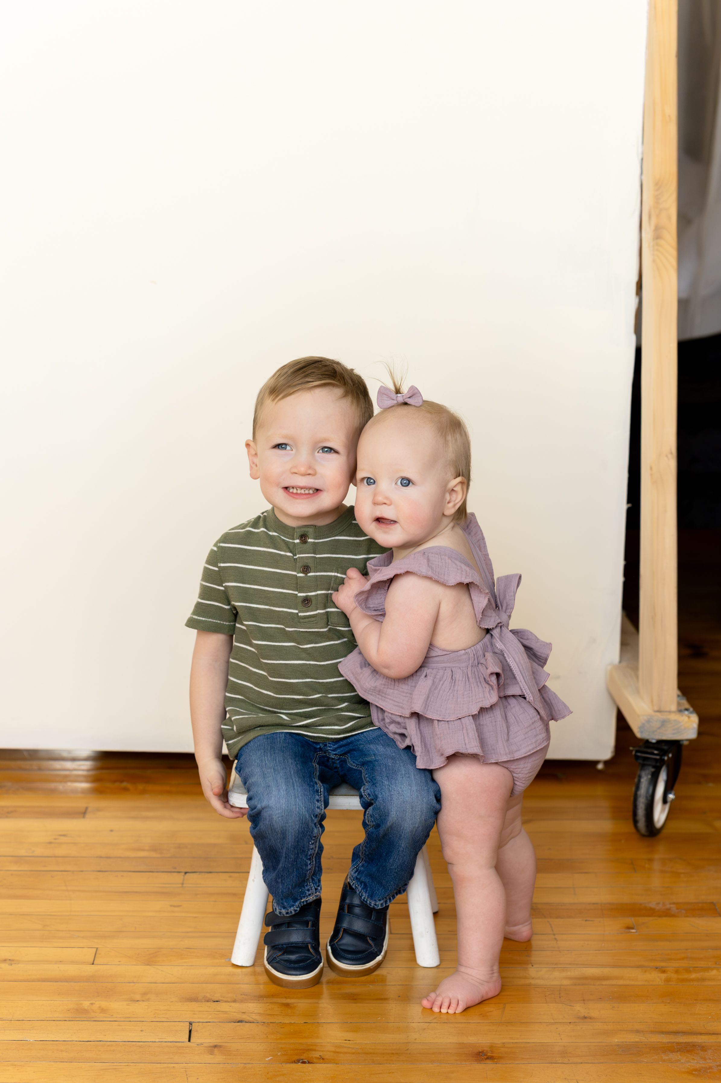 Two small children sit together on a wooden floor, one wearing a green striped shirt and jeans, the other in a purple dress.