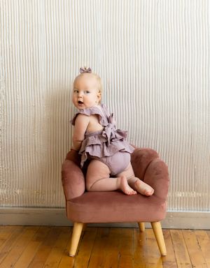 A small child in a ruffled romper sits on a brown upholstered chair with wooden legs against a striped wallpaper background.