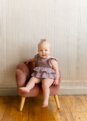 Small child in a ruffled dress sits on a modern brown leather chair against a striped wall.