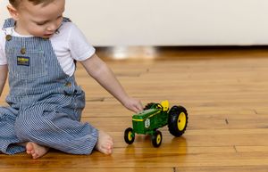 A toddler in striped overalls plays with a green John Deere toy tractor on a hardwood floor.