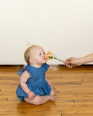 A baby in a blue dress sits on a hardwood floor reaching up towards a flower being held out.
