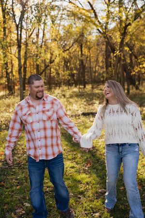 A couple in autumn woods wearing plaid shirt and white lace top hold hands while walking through golden leaves.