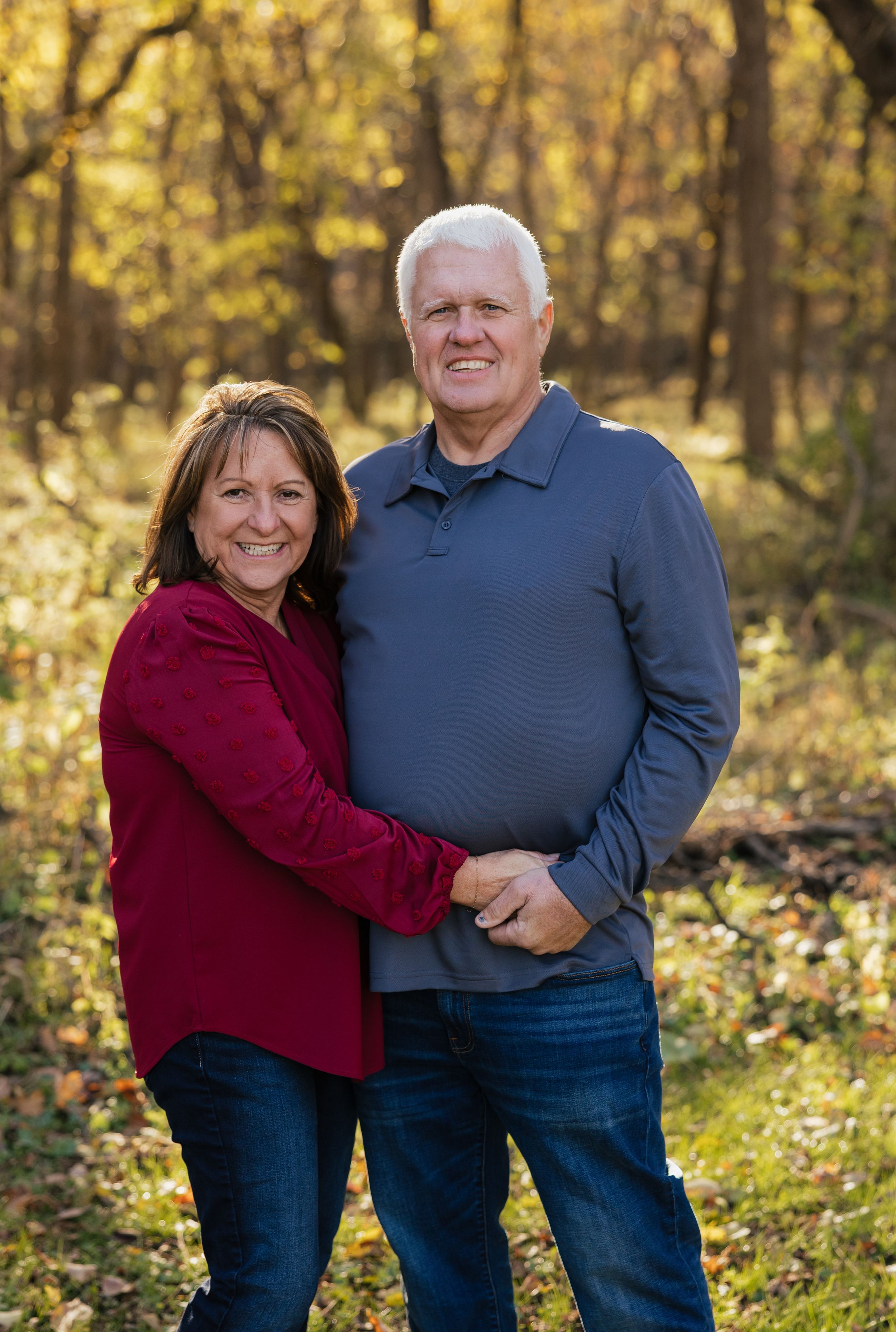 A couple shares loving embraces while posing outdoors in a wooded area with autumn foliage in the background.