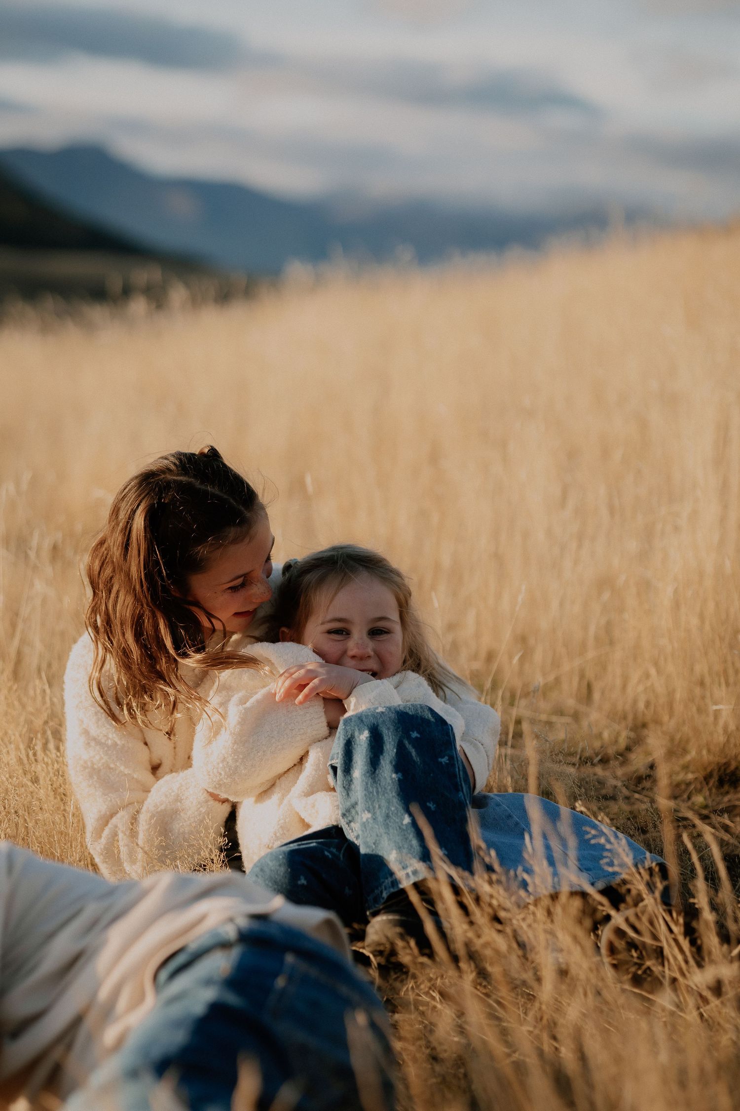 Black and white photo of a joyful young girl in a white cardigan playing in tall grass with mountains and lake behind her.
