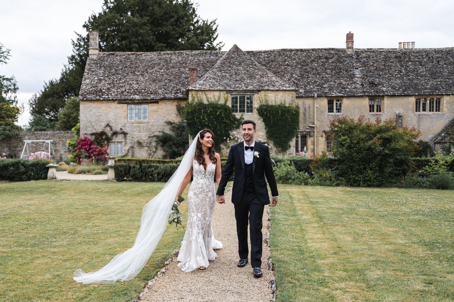 Bride and groom walking hand in hand on garden path before a historic Cotswolds stone manor house on their wedding day.