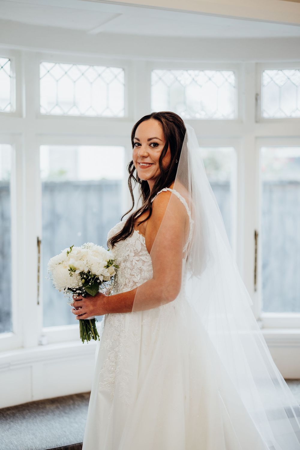 Bride in white lace gown and veil holds white bouquet, smiling in front of elegant leaded glass windows.