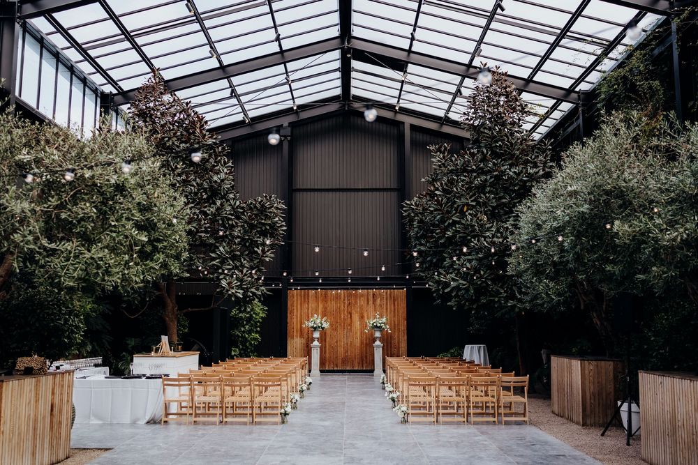 Elegant indoor wedding ceremony venue with wooden chairs, lush greenery, string lights, and a floral arch under a glass roof.