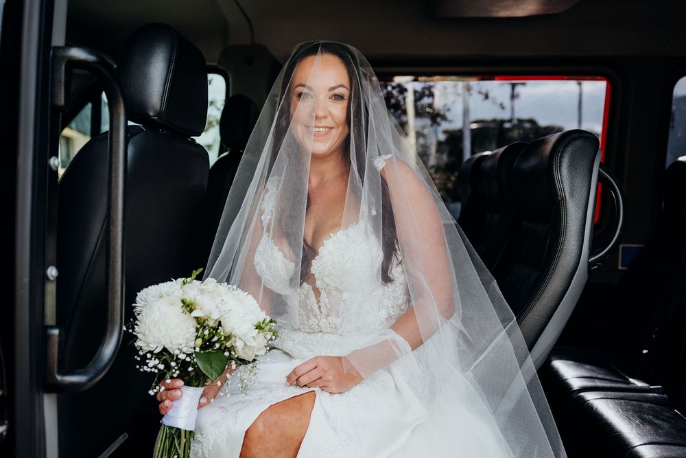 Bride in white lace gown and veil sits in vehicle holding white flower bouquet, smiling on wedding day.