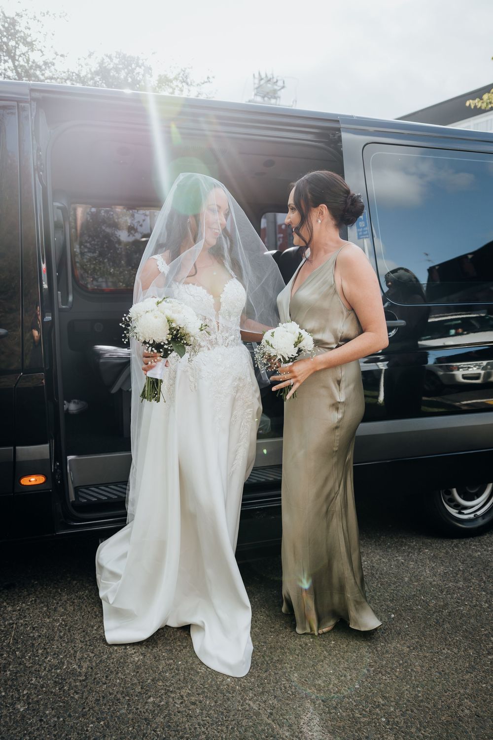 Bride in white lace gown and veil with bridesmaid in gold satin dress holding bouquets beside black van.