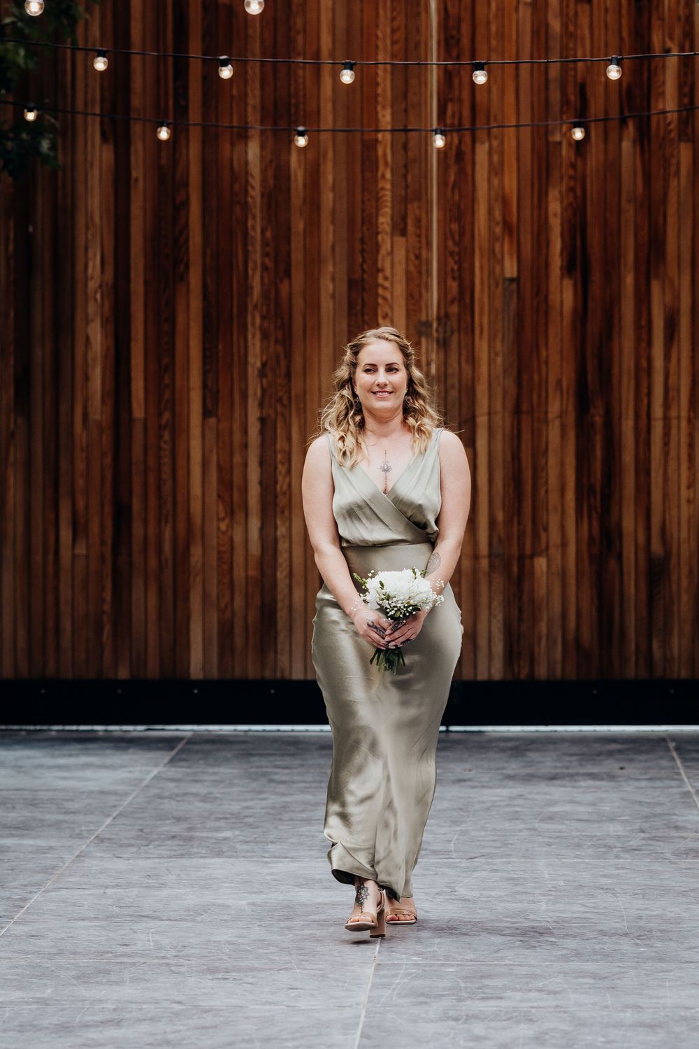 Bridesmaid in sage green satin dress holding white bouquet walking against rustic wooden wall with string lights.