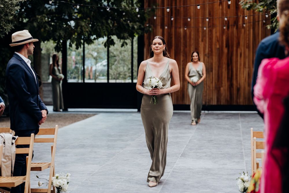 Bridesmaids in gold satin dresses walking down aisle at outdoor wedding ceremony with guests seated.