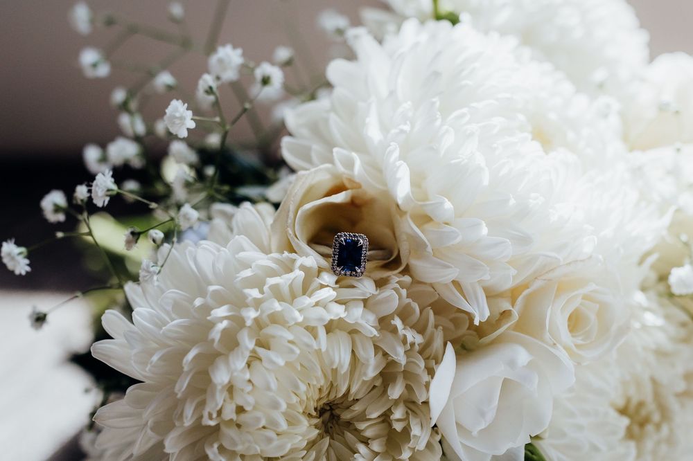 White chrysanthemum bridal bouquet with a sapphire and diamond engagement ring nestled among blooms.