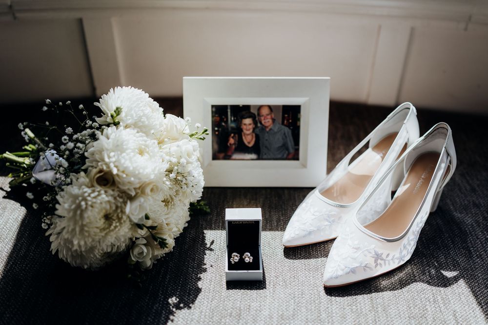 White wedding bouquet, lace heels, earring jewelry box, and couple photo frame arranged elegantly on a dark surface.