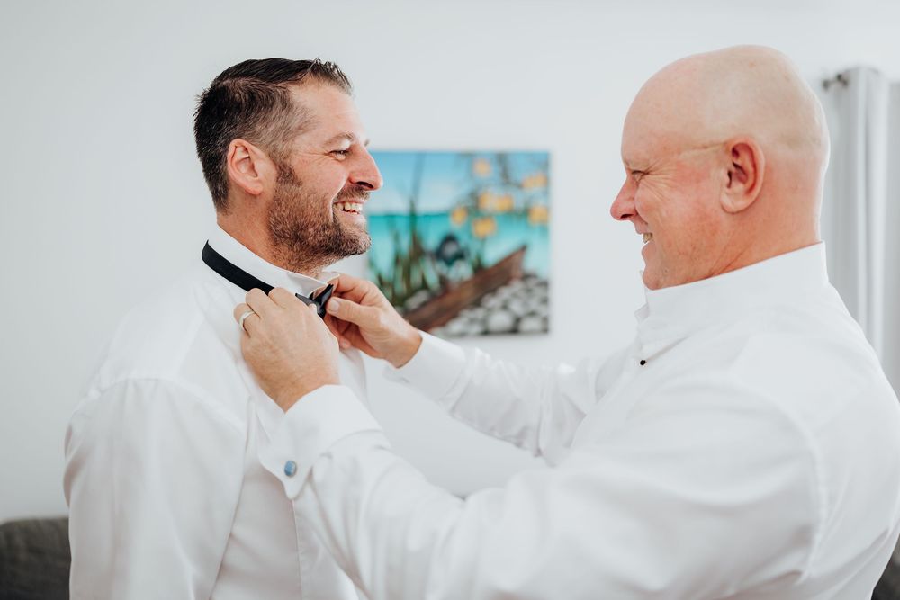 Father helping groom adjust bow tie while both smile during wedding day preparations.