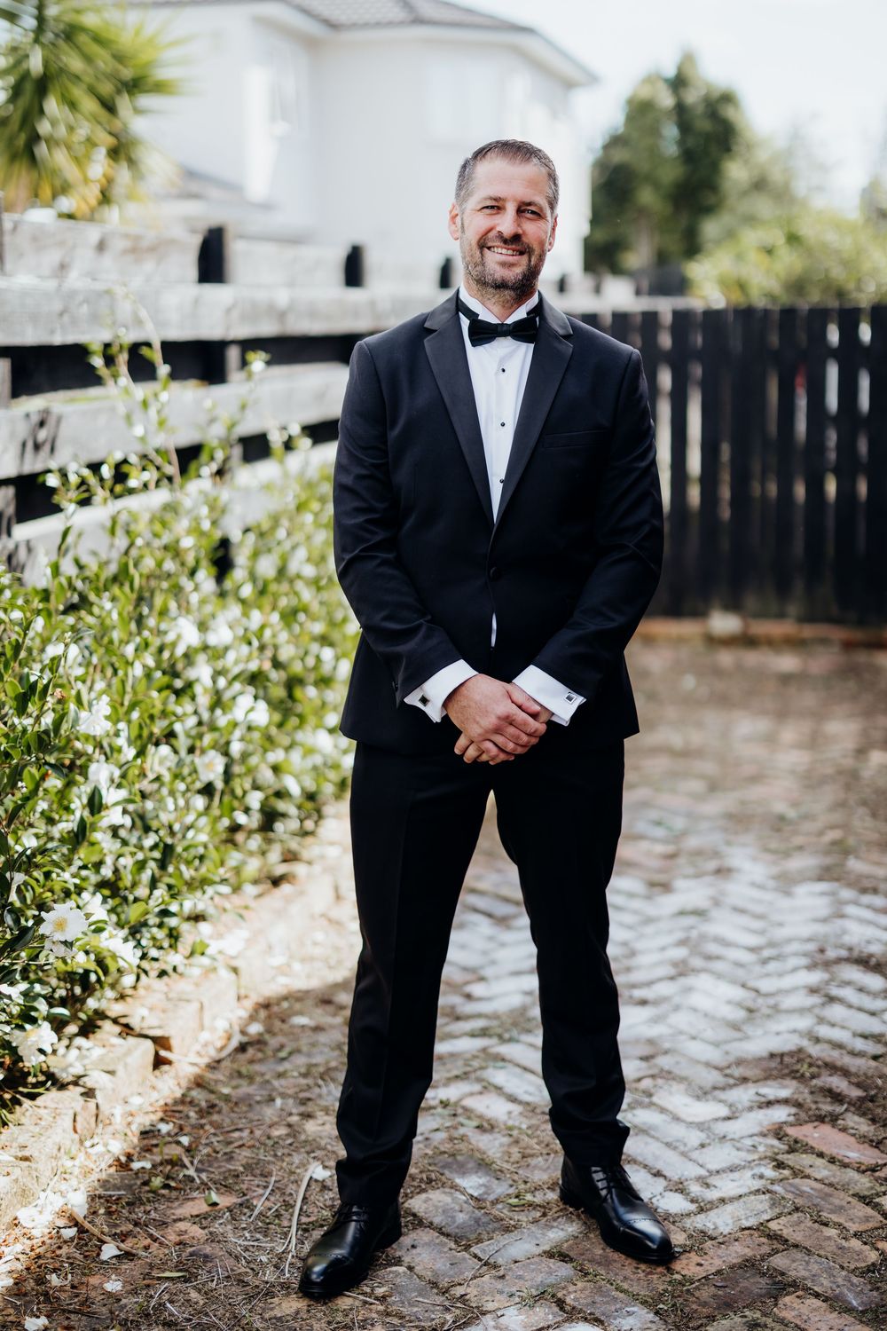 Smiling groom in black tuxedo and bow tie standing on brick path outdoors.